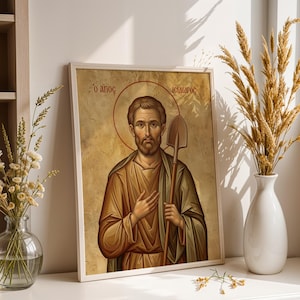 May include: Framed religious icon featuring a man with a halo, holding a shovel. The artwork is in warm tones of brown, beige, and gold, with Greek text at the top. The image is displayed on a white surface, next to a vase of dried wheat.