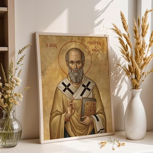 Op de afbeelding: Een religieus icoon in een lijst met een heilige met een halo, die een boek vasthoudt en gebaart. Het icoon heeft een goud- en beige kleurenpalet met Griekse tekst. De afbeelding wordt weergegeven op een wit oppervlak met decoratieve vazen.