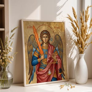 May include: Framed religious icon depicting a winged figure with a flaming sword and scales. The figure is clad in blue and gold armour, a red robe, and a halo. The background is gold with Greek text. The frame is white.