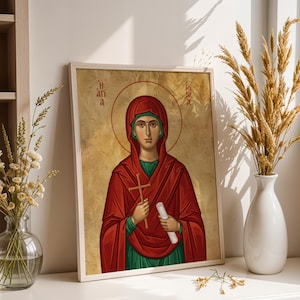 May include: A framed religious icon print featuring a figure in a red head covering and robe over a green garment, holding a wooden cross and a white scroll. The background is a textured gold color with a halo and Greek lettering in the upper corners.