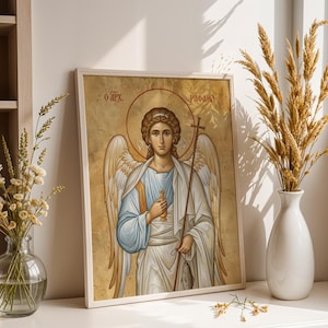 May include: Framed religious icon depicting an angel with a halo, holding a cross and a staff. The artwork has a gold background and is in a white frame. The text "O APX" and "PAФAHA" are visible. The image is displayed on a white surface.