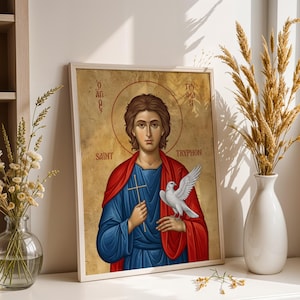 May include: A framed religious icon print featuring Saint Tryphon with brown hair, wearing a blue tunic and red cloak. The figure holds a small wooden cross and a white dove. The background is gold with Greek text and the English name Saint Tryphon.