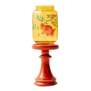 May include: A decorative, amber-colored glass jar with a goldfish illustration and floral accents, set atop a red, pedestal-style stand. The jar has a rectangular shape and is designed for display. The stand is a vibrant red color.