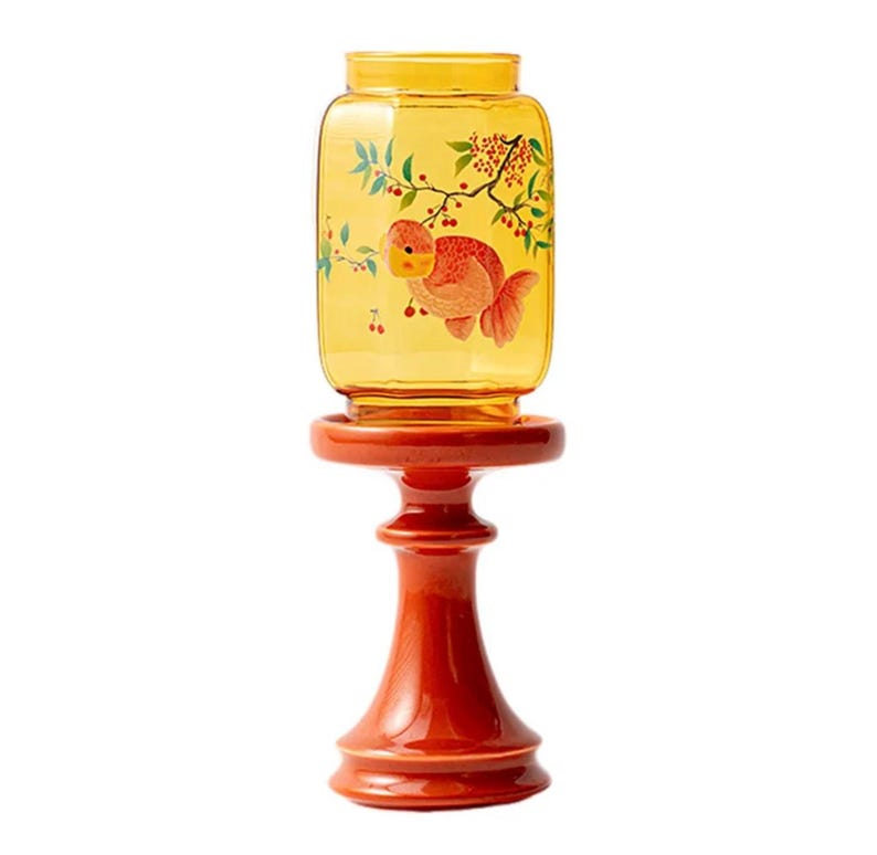 May include: A decorative, amber-colored glass jar with a goldfish illustration and floral accents, set atop a red, pedestal-style stand. The jar has a rectangular shape and is designed for display. The stand is a vibrant red color.