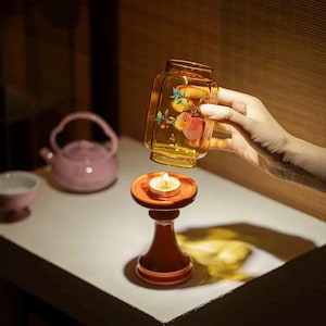 May include: A decorative glass jar with a fruit design is held above a small, orange candle holder with a lit candle. A pink teapot and teacup are in the background. The jar has a yellow tint and is decorated with a fruit and floral design.