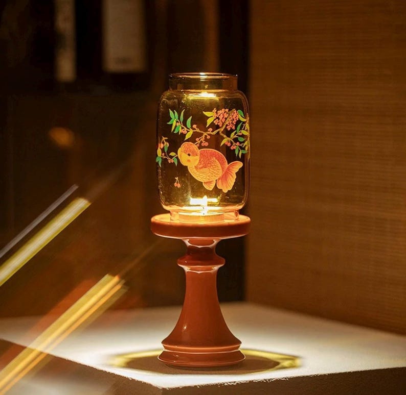 May include: A decorative glass jar with a goldfish and floral design, resting on a wooden stand. The jar is illuminated from within, creating a warm glow. The stand is a reddish-brown color.