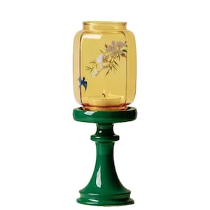 May include: A decorative candle holder featuring a yellow glass lantern with floral and bird designs, resting on a green pedestal. The lantern holds a lit candle, creating a warm glow. The design is elegant and suitable for home decor.