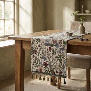 May include: A cream-coloured floral table runner with embroidered flowers in red, blue, and green. The runner has a green border and tassels. It is displayed on a wooden table. The table runner is approximately 180 cm in length.