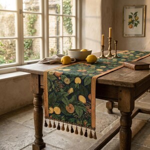 May include: A dark green table runner featuring a lemon and leaf design, with a tan border and tassels. The runner is displayed on a wooden table, alongside lemons, a bowl, and brass candlesticks. A framed botanical print is on the wall.