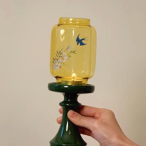May include: A yellow glass candle holder with a dark green base. The glass has a bird and floral design. The candle holder is held by a hand. The candle inside is lit.