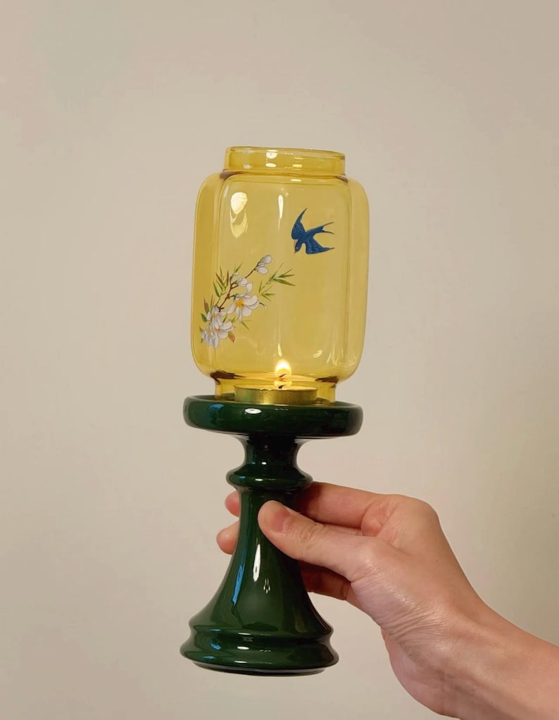 May include: A yellow glass candle holder with a dark green base. The glass has a bird and floral design. The candle holder is held by a hand. The candle inside is lit.
