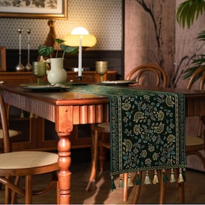 May include: A dark green table runner with a floral design and tassels. The runner is laid on a wooden dining table, with a lamp, candles, and other decorative items in the background. The runner features yellow, blue, and white floral elements.