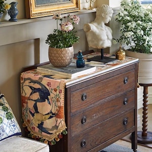 May include: A wooden chest of drawers with three drawers, adorned with a decorative runner depicting birds and floral motifs. A marble bust, potted plants, and a framed landscape painting enhance the room's aesthetic. A small blue bottle and books are also on display.