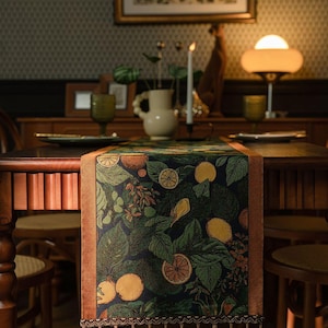 May include: A table runner with a dark green background featuring a botanical design of lemons and leaves. The runner has a brown border and is adorned with tassels. The runner is placed on a wooden table, creating a warm and inviting atmosphere.