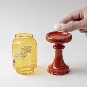 May include: A yellow glass jar with floral designs and a red candle holder. A hand is placing a small white candle on the candle holder. The candle holder is red and has a round top.