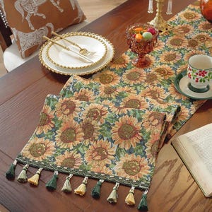 May include: A dining table arrangement with a sunflower-patterned table runner, adorned with tassels. Includes white plates with gold edging, gold cutlery, a teacup, and a glass bowl with sweets. A decorative cushion is in the background.