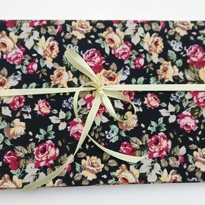 Könnte beinhalten: Ein Fotoalbum mit floralem Muster mit rosa und gelben Rosen auf schwarzem Hintergrund. Es ist mit einem hellgelben Band gebunden. Das Album hat eine Metallringbindung.