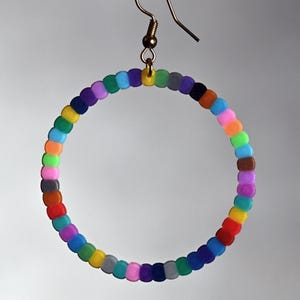 Könnte beinhalten: Ein einzelner Creolen-Ohrring mit einem goldfarbenen Haken. Der Reifen besteht aus kleinen, farbigen Perlen in einem Regenbogen von Farben, darunter Rot, Orange, Gelb, Grün, Blau und Lila. Der Ohrring hängt vor einem hellgrauen Hintergrund.