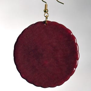 Könnte beinhalten: Ein einzelner, runder, weinroter Ohrring mit gewelltem Rand. Der Ohrring hängt an einem goldfarbenen Fischhaken. Der Ohrring hat eine einfarbige, glatte, glänzende Oberfläche.