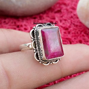 May include: A silver ring featuring a rectangular, faceted, deep pinkish-red gemstone set in a decorative metal frame with twisted rope detailing and scalloped edges. The text InduHandicraftHub is visible on the surface of the gemstone.