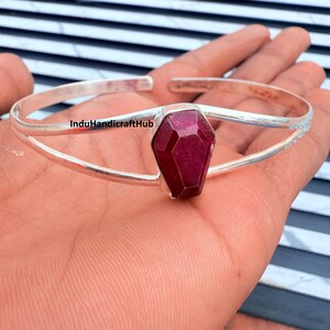Könnte beinhalten: Ein silbernes Armreif-Armband mit einem facettierten, tiefroten Edelstein. Der Edelstein ist der Mittelpunkt, gesetzt in einer geometrischen Form. Das Armband hat ein schlichtes, elegantes Design, perfekt für den Alltag.