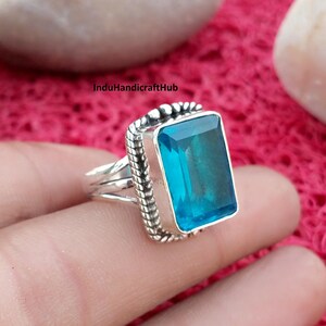 Puede incluir: Un anillo de plata de ley con una gran piedra preciosa rectangular facetada de color azul brillante. La piedra está engastada en un bisel con un borde de metal trenzado. La banda consta de tres hilos de plata finos y paralelos.