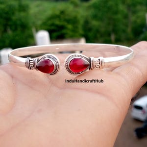 May include: A silver cuff bracelet featuring an open design with two teardrop-shaped red stones at the ends. The metal band displays intricate decorative detailing near the settings. The text InduHandicraftHub is visible on the bracelet.