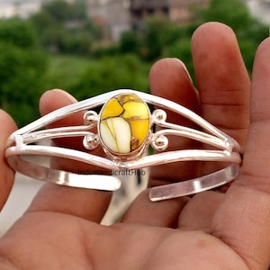 Puede incluir: Una pulsera de plata con una piedra central ovalada de color amarillo y blanco con vetas marrones. La banda de metal tiene un diseño dividido con detalles circulares decorativos a ambos lados de la piedra. El texto InduHandicraftHub es visible.