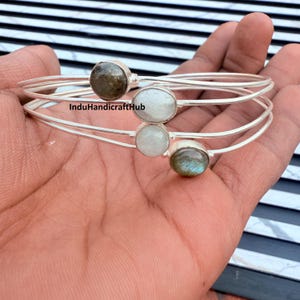 May include: Three silver-toned bangles, each adorned with a different gemstone: a dark green, a white, and a light green stone. The bangles are displayed on a hand. The text "InduHandicraftHub" is visible.