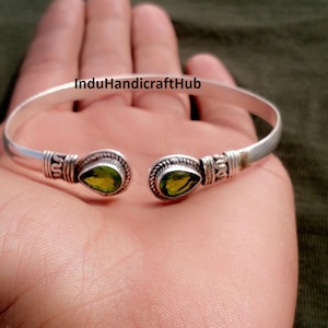 May include: A silver-toned cuff bracelet with two teardrop-shaped, light green gemstones. The bracelet has decorative detailing near the gemstones and the text "InduHandicraftHub" is visible.