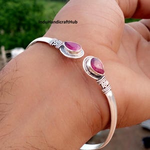 May include: A silver cuff bracelet featuring two teardrop-shaped pink gemstones at the open ends. The metal band displays intricate etched detailing and decorative wirework patterns surrounding each stone setting, creating a textured appearance.