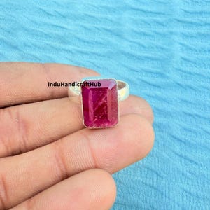 Może przedstawiać: Srebrny pierścionek z dużym, prostokątnym, głęboko różowym kamieniem szlachetnym. Pierścionek jest trzymany w dłoni na jasnoniebieskim tle. Na zdjęciu widoczny jest tekst "InduHandicraftHub".