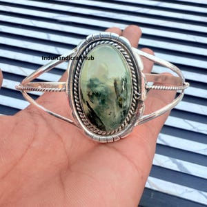 Peut inclure: Un bracelet manchette en argent orné d'une grande pierre précieuse ovale vert pâle avec des inclusions sombres rappelant de la mousse. La pierre est sertie dans un chaton torsadé, et le bracelet se compose de deux fins fils d'argent polis.