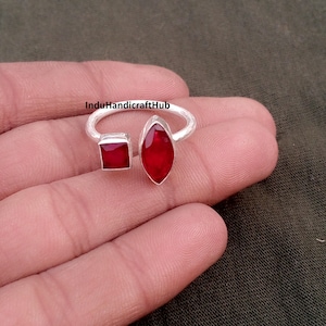 May include: A silver ring with two red gemstones: a square and a marquise-shaped stone. The open band design features the gemstones set on either side. The ring is shown on a hand.