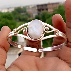 Pode incluir: Uma pulseira de prata tipo bracelete com uma pedra da lua oval central de superfície lisa e iridescente. A banda de metal apresenta detalhes decorativos de fio em espiral em ambos os lados da cravação. O texto InduHandicraftHub é visível.