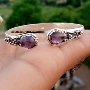 Può includere: Un bracciale a fascia in argento con due pietre di ametista viola a forma di goccia alle estremità aperte. La fascia in metallo presenta dettagli intricati in argento ossidato, inclusi piccoli accenti a perline e motivi a corda intrecciata.