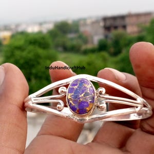 May include: A silver cuff bracelet featuring a central oval-shaped purple stone with gold-coloured veining. The metal band has a split design with decorative swirls on either side of the stone. The text InduHandicraftHub is visible on the band.