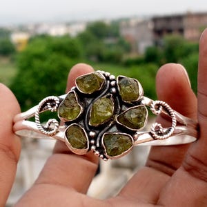 May include: A silver cuff bracelet featuring a central cluster of six raw, olive-green gemstones. The metal band displays a twisted rope texture on both sides of the stone setting, which is arranged in a circular, flower-like pattern.