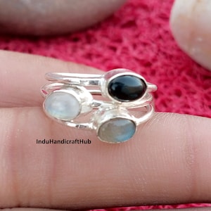 Può includere: Una pila di tre anelli in argento, ciascuno caratterizzato da una pietra preziosa liscia di forma ovale. L'anello superiore presenta una pietra nera, quello centrale una pietra bianca traslucida e quello inferiore una pietra grigio-blu. Il testo InduHandicraftHub è visibile.