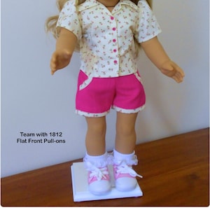 May include: A doll wearing a white short-sleeved shirt with a floral print and pink shorts. The doll is wearing white socks and pink sneakers. The text "Team with 1812 Flat Front Pull-ons" is visible below the doll.