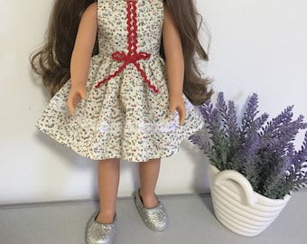 Puede incluir: Una muñeca con un vestido blanco con ribetes rojos y un lazo rojo. El vestido tiene un estampado floral. La muñeca tiene el pelo castaño y lleva zapatos plateados.