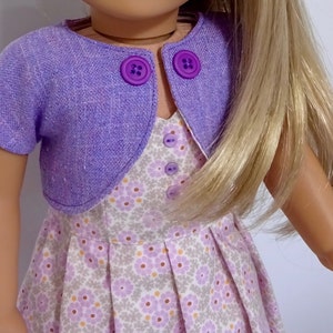 May include: A purple doll cardigan with two buttons on the front. The cardigan is worn over a white dress with a floral pattern. The doll has long blonde hair.