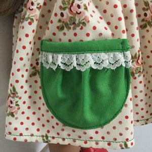 Può includere: Un vestito bianco con pois rossi e stampa floreale presenta una tasca verde con bordo in pizzo bianco.