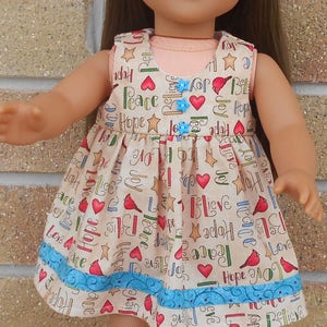 May include: A doll wearing a beige dress with a blue and white trim. The dress has a pattern of red hearts, stars, and the words "Hope", "Peace", "Love", and "Joy". The doll is wearing a white headband and black shoes.