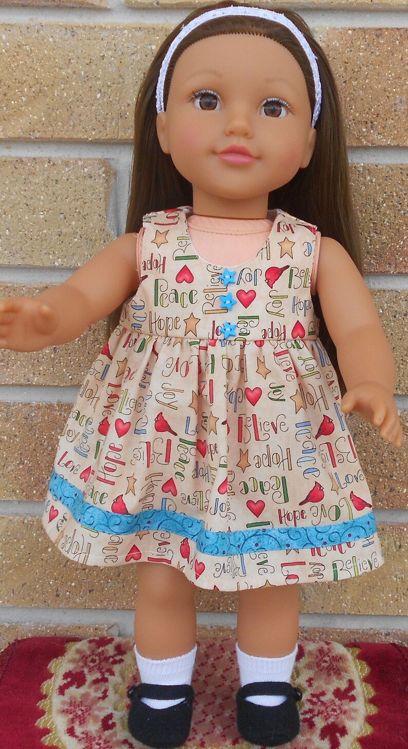 May include: A doll wearing a beige dress with a blue and white trim. The dress has a pattern of red hearts, stars, and the words "Hope", "Peace", "Love", and "Joy". The doll is wearing a white headband and black shoes.