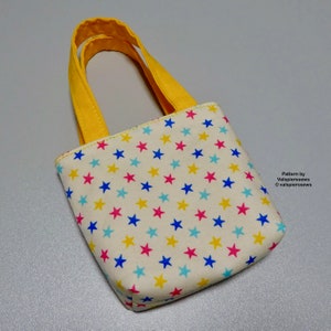 May include: A small, white fabric tote bag with yellow handles. The bag is covered in a pattern of blue, yellow, pink, and light blue stars. The text "Pattern by Valsgierssews ©valsgierssews" is printed on the bottom right corner of the bag.