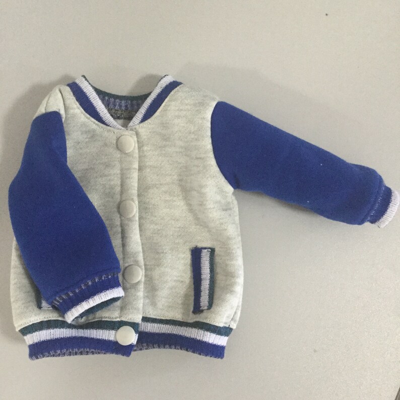 May include: A gray and blue baby jacket with a zipper closure and a pocket on the left side. The jacket has white and blue stripes around the cuffs and bottom.