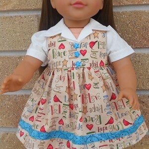 May include: A doll wearing a white collared shirt and a beige dress with a blue trim. The dress has a pattern of red hearts, stars, and birds with the words "Hope", "Love", "Joy", "Peace", and "Believe" repeated throughout.