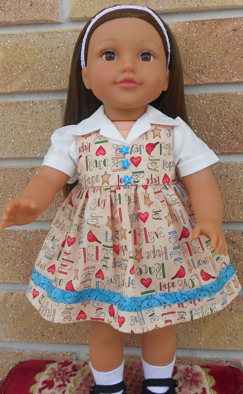 May include: A doll wearing a white collared shirt and a beige dress with a blue trim. The dress has a pattern of red hearts, stars, and birds with the words "Hope", "Love", "Joy", "Peace", and "Believe" repeated throughout.