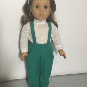 May include: A doll with brown hair and brown eyes wearing a white turtleneck shirt and green overalls. The doll has black shoes and a pink hair clip. The doll is standing in front of a white background.